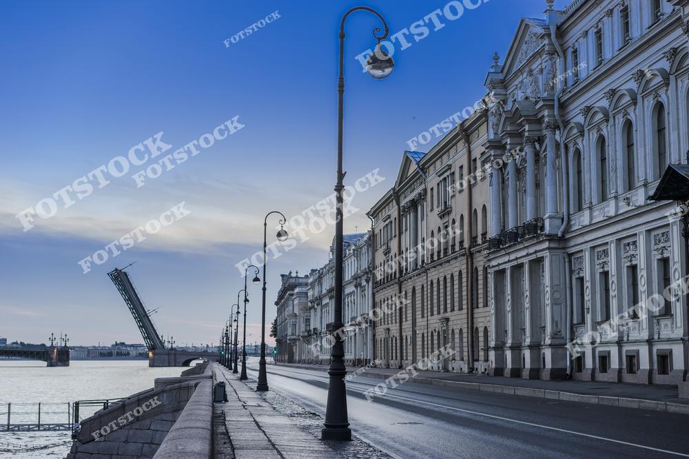На фотографии изображена набережная Невы в Санкт-Петербурге на рассвете. Разведённый мост вдалеке символизирует начало нового дня, а пустынная улица и старинные здания на переднем плане подчёркивают исторический характер этого района.